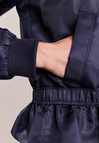 Chaqueta azul marino con una textura suave y brillante; presenta una cinturilla y puños elásticos, detalles fruncidos y un bolsillo lateral.