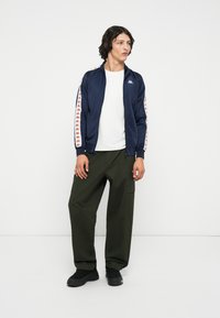 Mornarica zip-up jakna s bijelim i narančastim bočnim trakama, nosi se preko bijele majice; kombinira se s olivno zelenim slobodnim cargo hlačama i crnim cipelama.