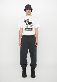 Weißes Baumwoll-T-Shirt mit einem schwarzen Grafiken eines Hundes und dem Text "CERBERUS." Graue Sweatpants mit seitlichem Logo-Print, kombiniert mit schwarzen Stiefeln und einer Mütze.