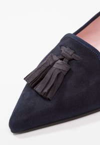 Mocassin pointu en daim bleu marine avec détail de pompon en daim noir et doublure intérieure rose, présenté sur un fond blanc.