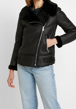 Femme portant une veste en cuir noire avec col et poignets en fourrure, fermeture éclair asymétrique, associée à un jean bleu clair.