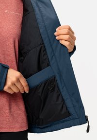 Marineblaue Outdoor-Jacke mit glatter Oberfläche, sichtbarer Innenfutter und einer Reißverschlusstasche. Mit einem Stoff-Patch mit der Aufschrift "RE:CYCL."