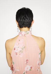Personne aux cheveux courts noirs porte une robe rose à fleurs à col licou et des boucles d’oreilles pendantes transparentes, tournée dos à la caméra.