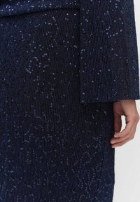 Laivansinis mekko, jossa on pystysuuntainen ribbitekstuuri, koristeltu paljeteilla, pitkät hihat ja sulavalinjainen siluetti.