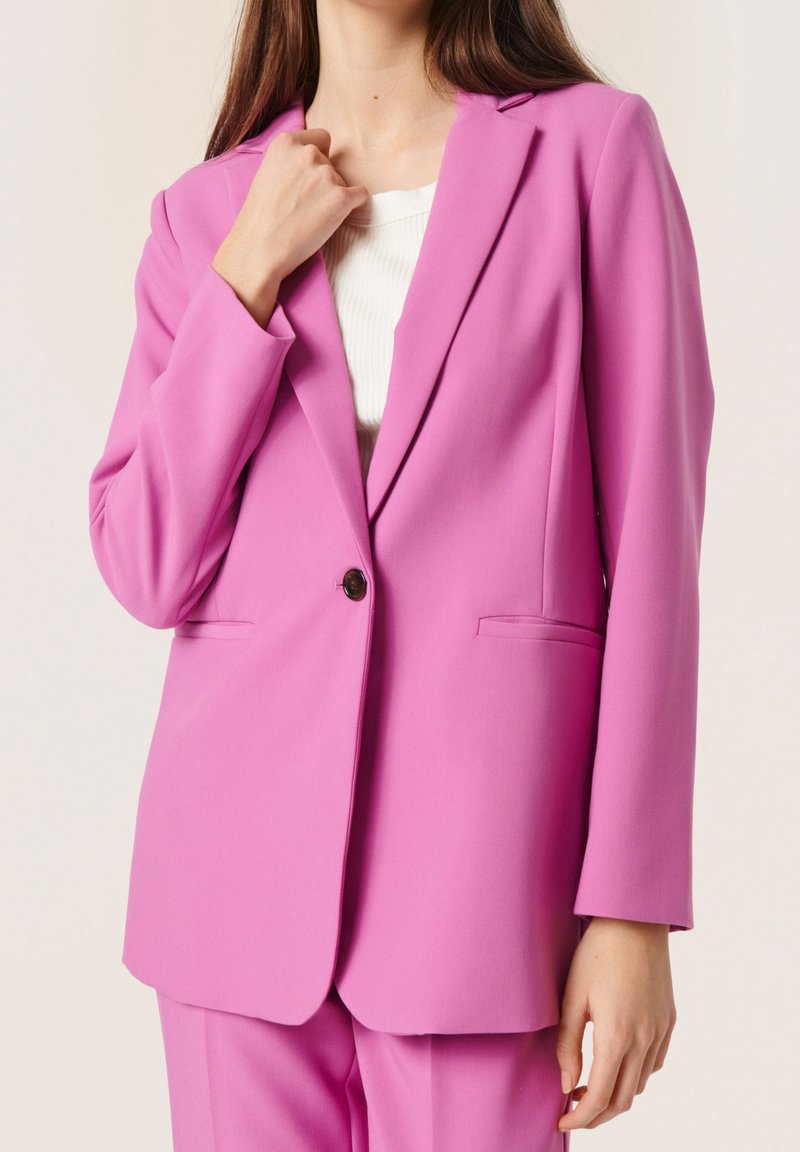 Růžový blazer s jedním knoflíkem, klopami ve tvaru výkroje, předními kapsami a strukturovaným designem. Nositý přes bílý texturovaný top.