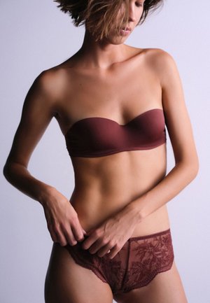 Femme ajustant sa culotte en dentelle, portant un soutien-gorge sans bretelles, avec des cheveux courts et décoiffés, sur un fond clair uni.