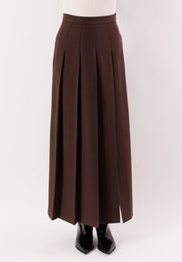 Gonna lunga plissettata marrone indossata con top bianco e stivali neri con punta affilata, mostrata su uno sfondo bianco pulito.