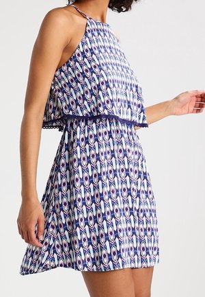 Frau in ärmellosem, über dem Knie endendem Kleid mit blauem, weißem und lila geometrischem Muster und geschichtetem Oberteil vor einfarbigem Hintergrund.