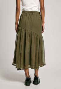 Jupe midi en olive, à étages, fabriquée en tissu léger avec une taille élastique et un ourlet froncé. Des chaussures à plateforme noires brillantes sont visibles.