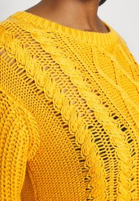 Pull jaune tricoté avec un motif texturé, comprenant un design à torsades et des sections en maille ajourée, col rond.