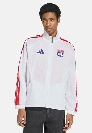 Giovane uomo che indossa una giacca sportiva bianca con zip, con strisce rosse sulle maniche e i loghi di Olympique Lyonnais e Adidas sul petto.
