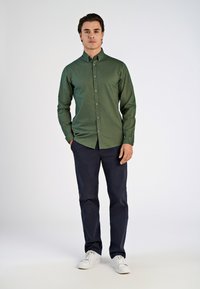 Camisa verde de botones, hecha de un tejido texturizado, de mangas largas y con un diseño ajustado. Combinada con pantalones azul marino y zapatillas blancas.