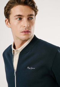 Mornarica majica na zatvaranje sa ribanim okovratnikom, sa svijetlom majicom polo ispod i izvezenim logom na prsima.