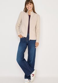 Beige takki, jossa on fleece-vuori, ribbaustekstuuri ja korkeahko kaulus, yhdistettynä tummansinisiin leveälahkeisiin farkkuihin ja valkoisiin lenkkareihin, joissa on punaisia yksityiskohtia.