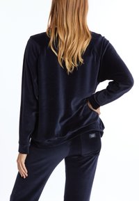 Navyblauwe velours lounge-uitrusting, met lange mouwen en een ontspannen pasvorm. Duidelijke textuur, met een klein merklabel op de achterkant van de broek.