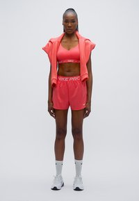 Ροζ αθλητικό σουτιέν και σορτς Nike Pro, συνδυασμένα με ροζ φούτερ με κουκούλα που είναι περασμένο στους ώμους. Λευκά αθλητικά παπούτσια και λευκές κάλτσες ολοκληρώνουν την εμφάνιση.