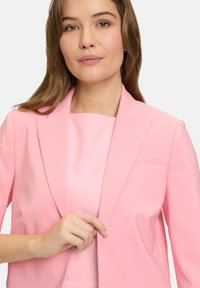 Jasnoróżowa marynarka o dopasowanym kroju z wysuniętymi klapami i jedną kieszenią na piersi. Gładki, usztywniony materiał. Model trzyma marynarkę lekko otwartą.