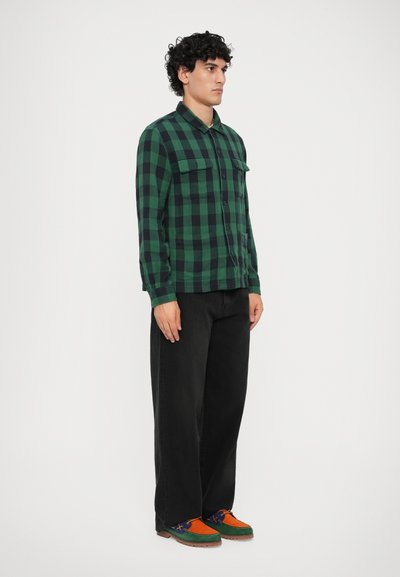 Camisa de flanela xadrez verde e preta com fecho de botão na frente, duas Bolsos no peito, combinada com calças largas pretas e sapatos multicoloridos.