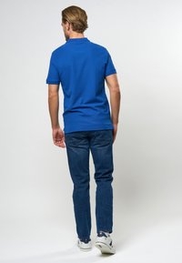 Homme aux cheveux châtain clair portant un polo bleu, un jean bleu et des baskets blanches, debout de dos sur un fond uni.
