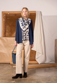 Tmavomodrý kardigán cez krémovú blúzku s potlačou, doplnený krémovými nohavicami a hnedej členkovej členkovej obuvi na opätku. Súčasťou je modrá kabelka na rameno.