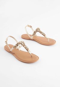 Guldtonade platta sandaler med dekorerade remmar, beige fotbädd och justerbar ankelförslutning. Slimmad design, öppen tå-stil och minimalistisk silhuett.