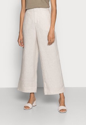 Femme portant un pantalon large en lin beige et des sandales à talons blocs ouvertes tressées, se tenant devant un fond uni.