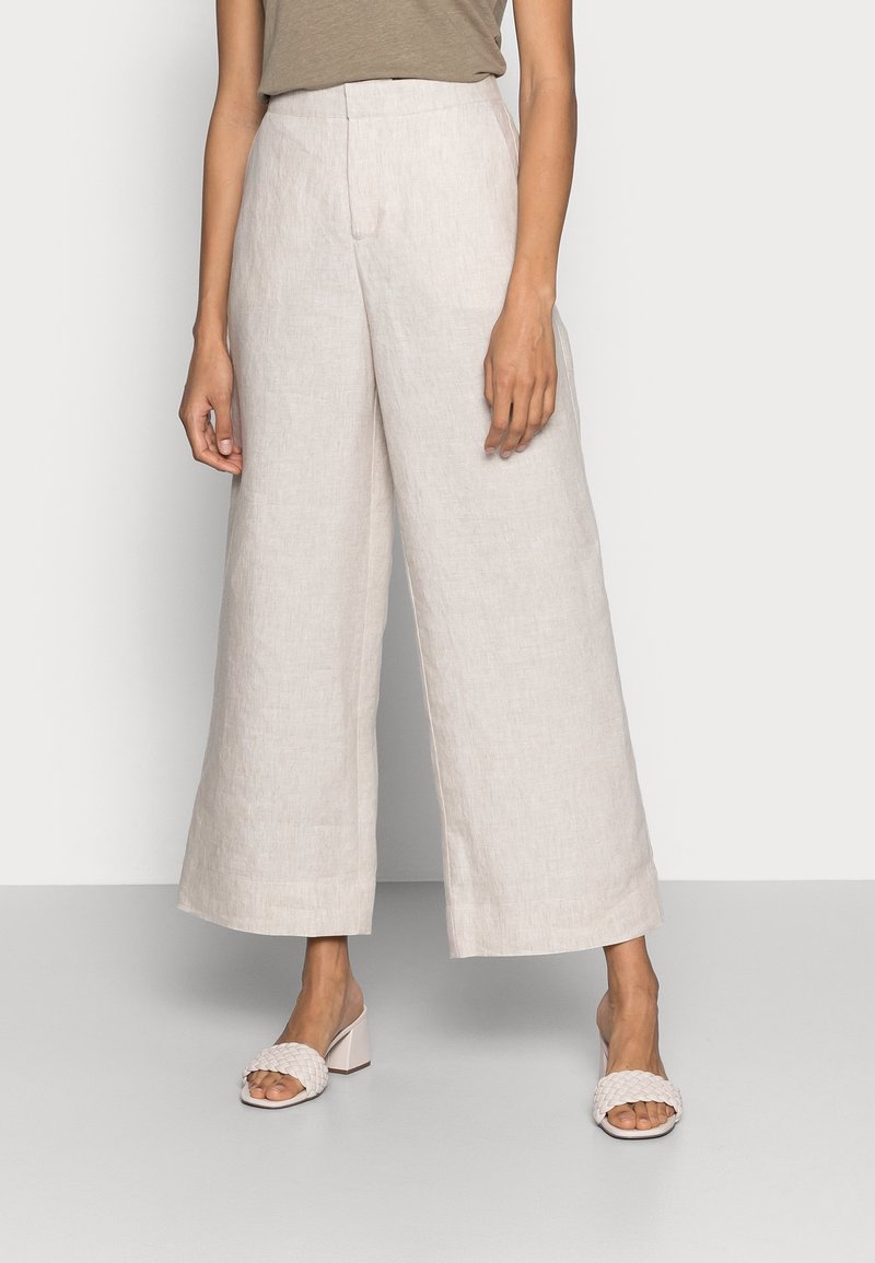Femme portant un pantalon large en lin beige et des sandales à talons blocs ouvertes tressées, se tenant devant un fond uni.