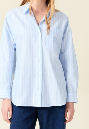 Frau trägt ein hellblau gestreiftes Hemd mit Knöpfen, einer Brusttasche und dunkelblaue Hosen, steht vor einem schlichten Hintergrund.