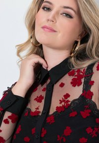 Camicia nera trasparente con ricamo floreale rosso, dettagli in pizzo e collo a camicia. Presenta bottoni lungo il davanti e maniche lunghe.