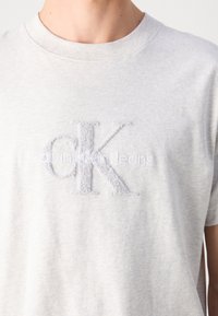 Primer plano de una camiseta gris claro con el logo texturizado "CK" y el logo bordado "Calvin Klein Jeans" en el pecho.