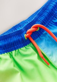 Blue and green swim shorts with an elastic waistband, featuring a bright orange drawstring and smooth fabric texture.