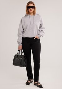 Nő szürke kapucnis pulóverben, fekete nadrágban, fekete szegecses cipőben, nagy fekete napszemüvegben, és fekete kézitáskát tart egy egyszínű háttér előtt.