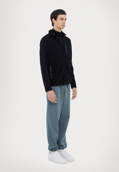 Vesta din fleece neagră cu guler înalt și buzunar cu fermoar, asortată cu pantaloni de trening lejeri de culoare albastru deschis și adidași albi. Textură plată.