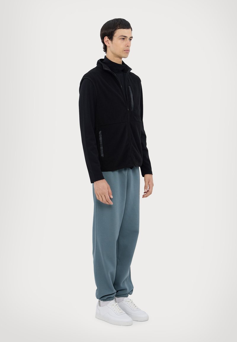 Gilet en polaire noir avec col montant et poche zippée, associé à un pantalon de jogging bleu clair ample et des baskets blanches. Texture plate.