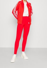 Červená sportovní bunda s bílými pruhy a odpovídajícími červenými legínami. Obě mají logo Adidas. Doplněno bílými teniskami.
