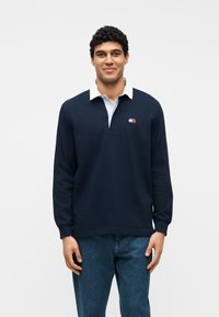 Άνδρας με σγουρά σκούρα μαλλιά που φοράει μπλε μακρυμάνικο πόλο μπλουζάκι με λευκό γιακά και μπλε τζιν, στέκεται μπροστά σε λευκό φόντο.