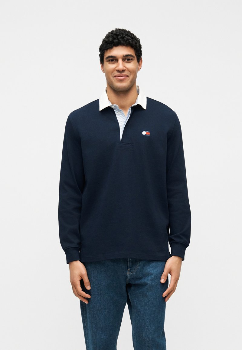 Άνδρας με σγουρά σκούρα μαλλιά που φοράει μπλε μακρυμάνικο πόλο μπλουζάκι με λευκό γιακά και μπλε τζιν, στέκεται μπροστά σε λευκό φόντο.