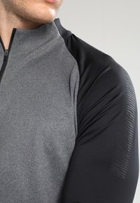 Camiseta atlética de manga corta con un cuerpo gris y mangas negras, fabricada en tejido suave, incluye una cremallera frontal y acentos texturizados en los hombros.