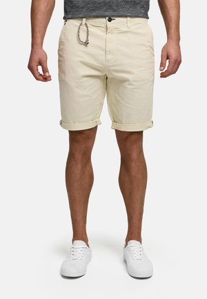 Mies, joka pukeutunut beigeihin käännettyhelmaisiin shortseihin, joissa on nappi ja köysidetalji, yhdistettynä valkoisiin tennareihin ja harmaaseen t-paitaan.