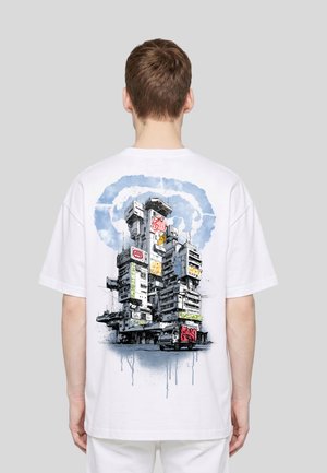 Personne portant un t-shirt blanc avec une illustration détaillée d'un bâtiment urbain futuriste à plusieurs niveaux et un camion à l'avant.