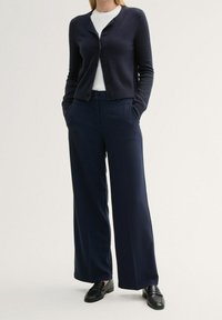 Navy Cardigan mit Knöpfen, strukturiertem Strick, über einem weißen Rippenshirt getragen. Kombiniert mit weiten navy Hosen und schwarzen Slippern.