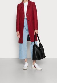 Abrigo rojo de mezcla de lana con solapa en pico y dos botones, llevado sobre una blusa blanca y jeans de pierna ancha azul claro. Bolsa de mano de cuero negro, zapatillas blancas.