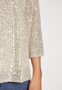 Haut à manches courtes scintillant recouvert de sequins dorés et argentés, avec un tissu léger et texturé, présentant un ourlet arrondi.