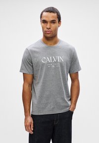 Mężczyzna w szarej koszulce Calvin Klein i ciemnych dżinsach, stojący z jedną ręką w kieszeni na tle białym.