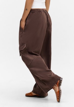 Person iført løse brune cargobukser med sidelommer og brune sandaler med remme, stående med det ene ben bøjet.