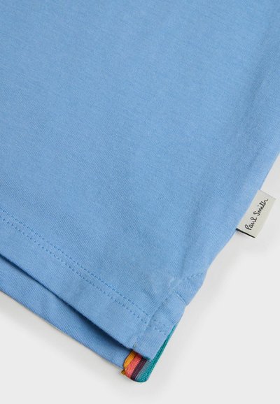 Ljusblå bomullströja med korta ärmar, med en sydd fåll och en etikett. Accenter av grönt, rött och gult vid nederkanten.
