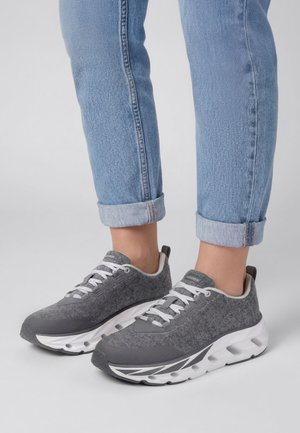 Personne portant un jean bleu clair roulé aux chevilles et des baskets de sport grises avec des semelles blanches épaisses sur un fond clair uni.
