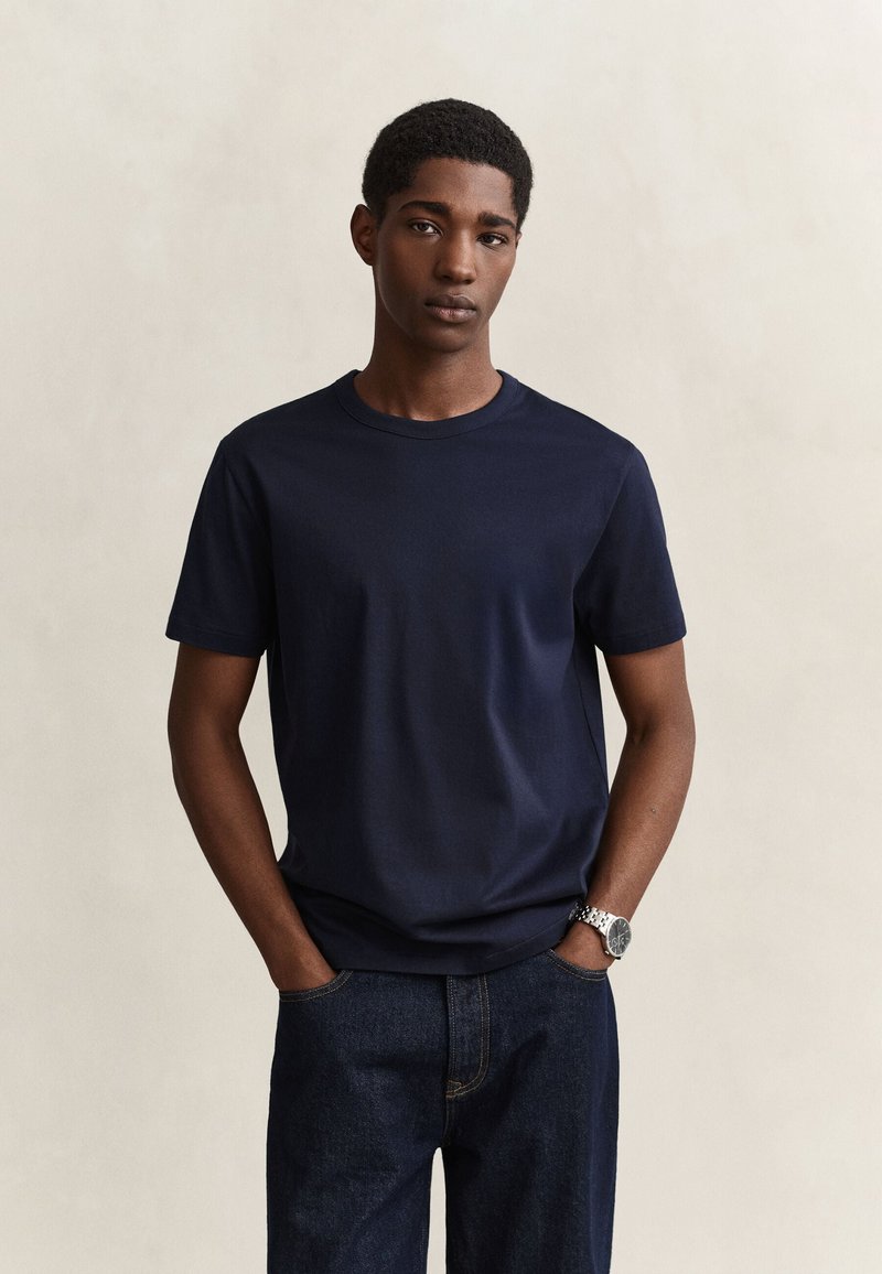 Jeune homme aux cheveux courts et bouclés, portant un t-shirt bleu foncé et un jean, debout les mains dans les poches devant un fond uni.