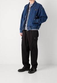 Blaue Denim-Bomberjacke mit Reißverschluss, elastischem Bund und Logo-Patch. Kombiniert mit schwarzen Cargo-Hosen und schwarzen Schuhen.