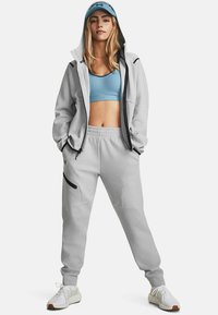 Haut de sport gris clair avec des accents noirs, porté sur un soutien-gorge de sport bleu et un pantalon de survêtement gris assorti. Baskets blanches avec des détails texturés.